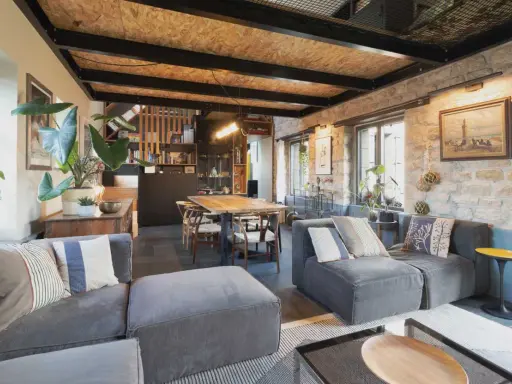 Salon cosy d'une maison de pêcheur rénovée, avec canapés gris, table en bois, murs en pierre et plantes vertes.