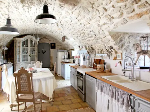 Cuisine rustique avec des vieux meubles patinés, un buffet ancien, une table dressée et des murs en pierre.