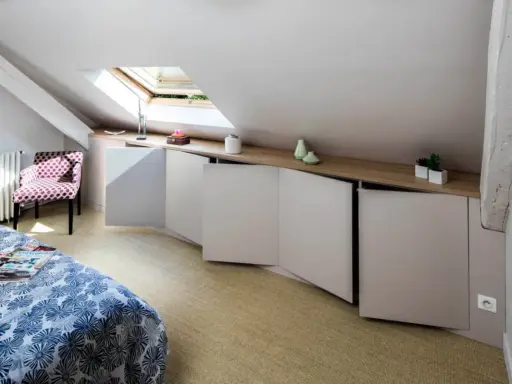 Chambre mansardée d'un coté avec lit, fauteuil à pois et rangements bas sur mesure. Un velux éclaire la pièce.