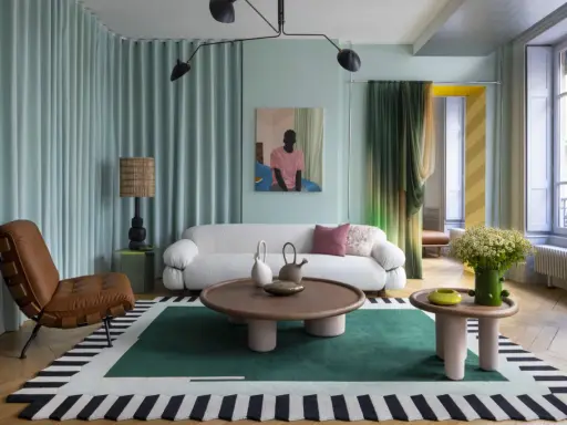 Un salon moderne avec un canapé blanc bouclé, un fauteuil en cuir marron, une table basse ronde et un tapis graphique. Une décoration appartement audacieuse et colorée.