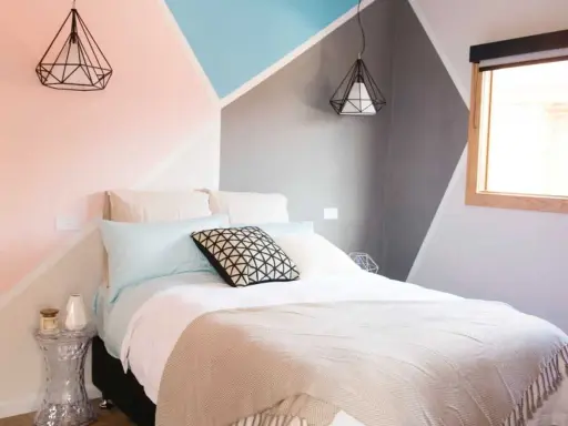 Lit moderne avec une tête de lit à peindre aux formes géométriques roses, bleues et grises. Deux suspensions noires en forme de diamant surplombent le lit.