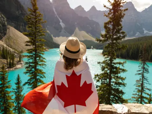 Vue d'une femme avec un drapeau canadien sur les épaules, contemplant un lac turquoise et des montagnes. Un rêve de déménagement France Canada.