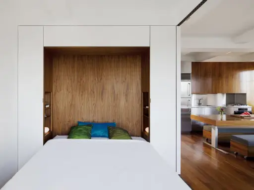 Un petit espace chambre salon optimisé avec un lit escamotable en bois, des coussins colorés et une cuisine moderne adjacente.