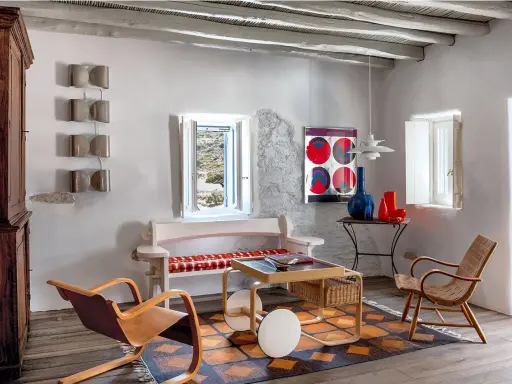 Salon d'une typique maison grecque avec mobilier en bois, tapis coloré et vue sur la mer par la fenêtre.