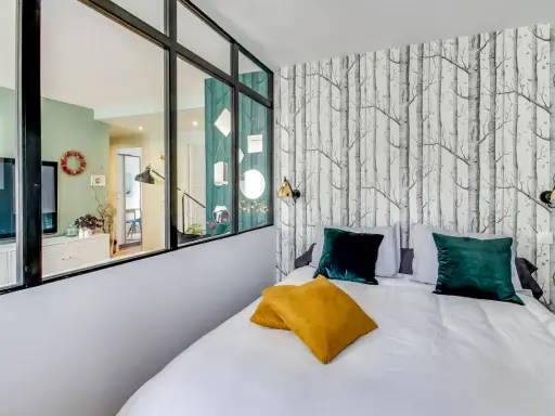 Idée pour comment amenager une chambre en longueur : un lit confortable avec des coussins colorés, un papier peint forêt et une verrière pour séparer les espaces.