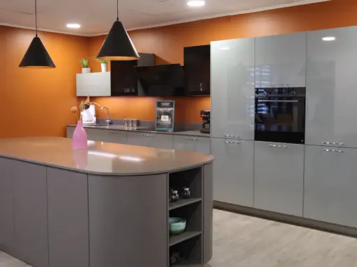 Un exemple de cuisine moderne aux lignes épurées, avec un îlot central gris et des meubles hauts gris clair. L'orange vif du mur apporte une touche de dynamisme.