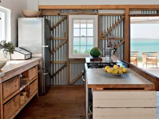 Cuisine campagne chic avec réfrigérateur, plan de travail en bois brut, murs en tôle ondulée et vue sur la mer.