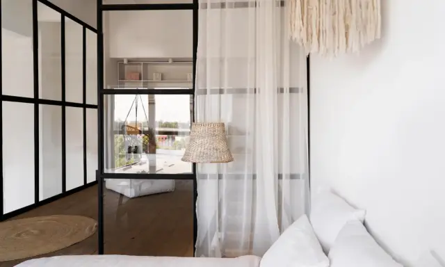 Verrière noire séparant une chambre en deux, avec une seule fenêtre visible. Un rideau blanc et un abat-jour en rotin ajoutent une touche cosy.