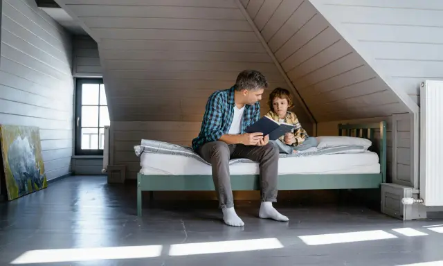 Père lisant une histoire à son fils sur un lit sous pente, dans une chambre mansardée.