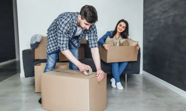 Un homme apprend comment scotcher un carton pour déménager, tandis qu'une femme assise sur un canapé tient une autre boîte remplie d'objets.