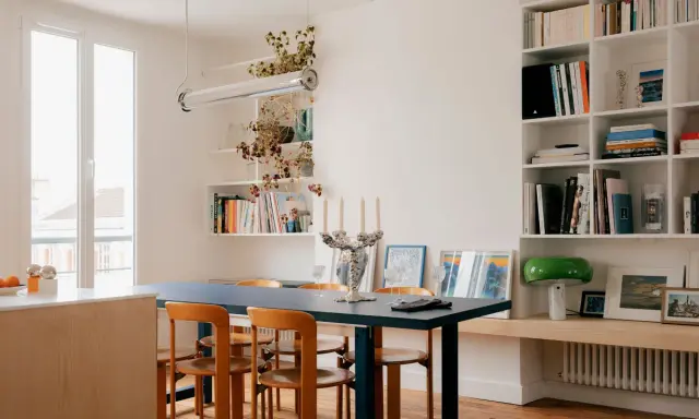 Salle à manger lumineuse d'un appartement traversant, avec table bleue, chaises en bois et étagères remplies de livres.