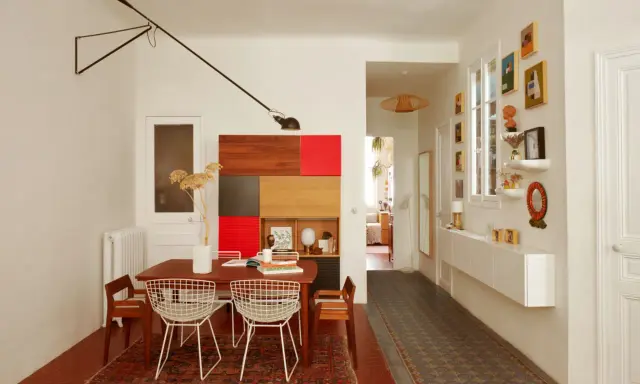 Salle à manger et couloir d'une maison ancienne, avec une table en bois, des chaises en fil de fer, une étagère colorée et des tableaux.