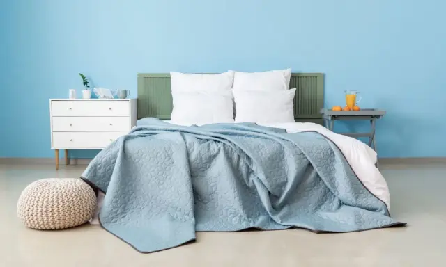 Une chambre paisible avec un lit douillet recouvert d'une couverture bleu glacier. Un pouf moelleux et une commode blanche complètent l'ensemble.