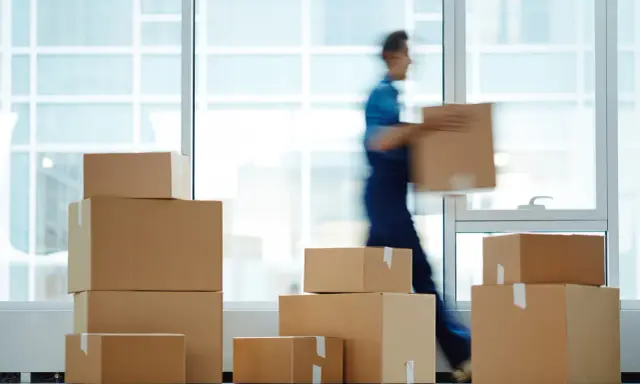 Un déménageur porte une boîte, entouré de cartons empilés. Une scène typique pour une facture déménagement.