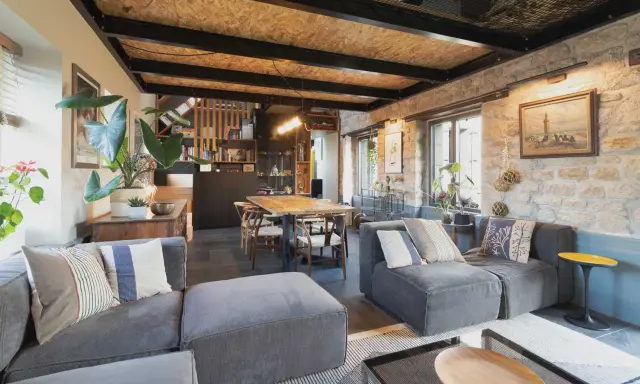 Salon cosy d'une maison de pêcheur rénovée, avec canapés gris, table en bois, murs en pierre et plantes vertes.