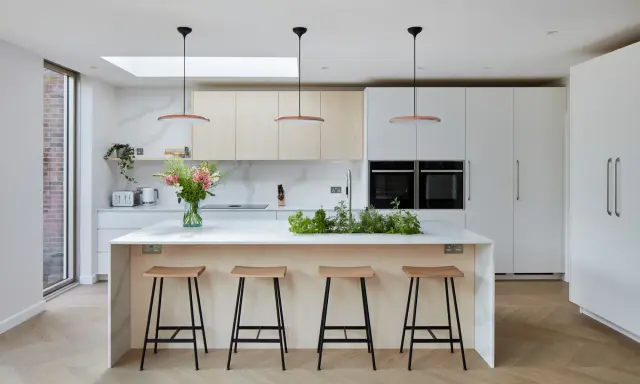 Élégante cuisine ouverte avec îlot central, tabourets en bois, et un jardin d'herbes aromatiques. Des armoires blanches et des fours intégrés complètent ce design moderne.
