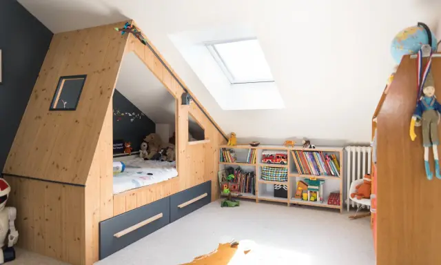 Chambre d'enfant aménagée sous les combles avec un lit cabane en bois, étagères remplies de jouets et livres. Un plan d'aménagement astucieux pour optimiser l'espace.