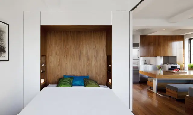 Un petit espace chambre salon optimisé avec un lit escamotable en bois, des coussins colorés et une cuisine moderne adjacente.