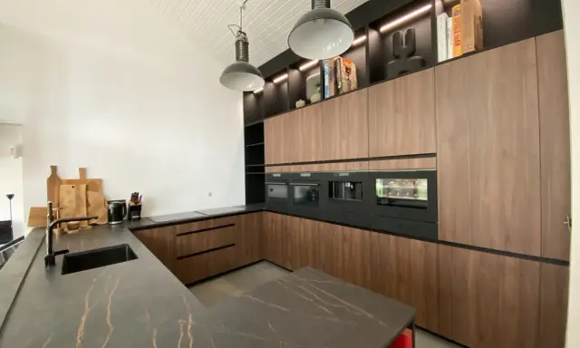 Cuisine moderne avec îlot central, plan de travail en marbre noir veiné, meubles en bois, électroménagers intégrés. L'implantation cuisine est pensée pour l'efficacité.