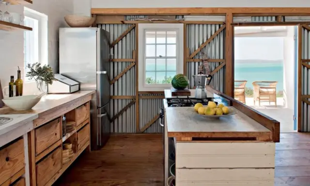 Cuisine campagne chic avec réfrigérateur, plan de travail en bois brut, murs en tôle ondulée et vue sur la mer.