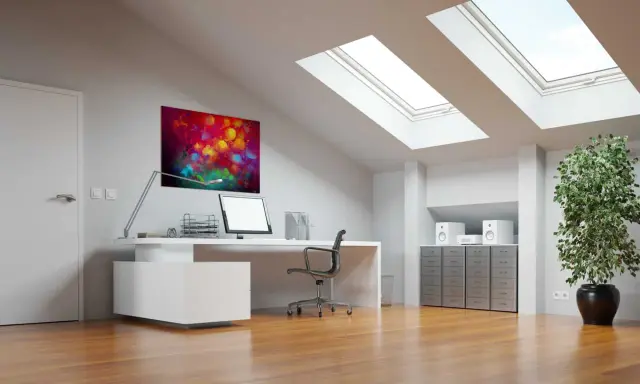 Bureau sous pente lumineux avec tableau abstrait coloré, ordinateur, plante verte et puits de lumière.