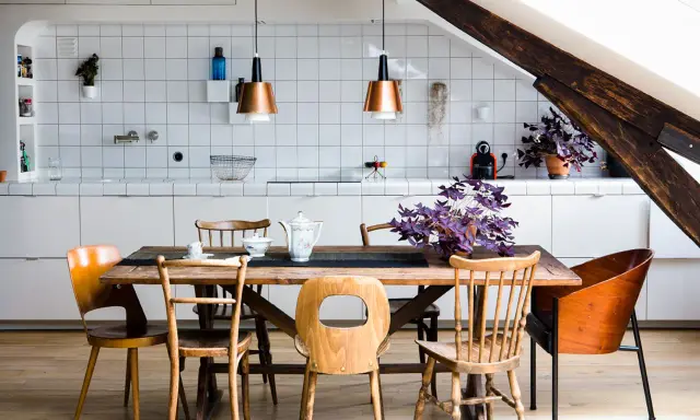 Espace de vie lumineux avec cuisine, salle à manger et salon dans la même pièce. Table en bois entourée de chaises, murs carrelés blancs, poutre de bois apparente.