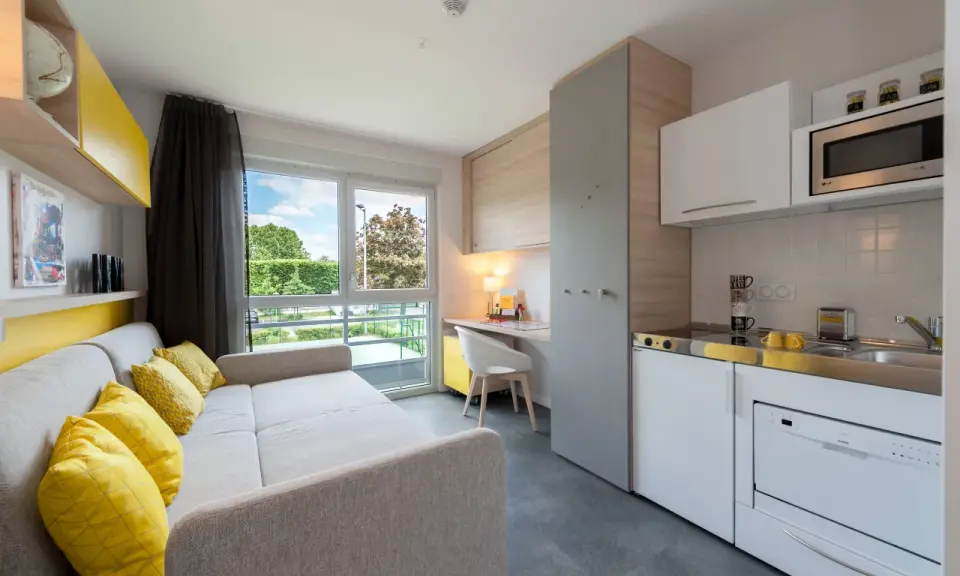 Aménagement studio moderne avec canapé, coin cuisine, bureau et vue sur jardin.