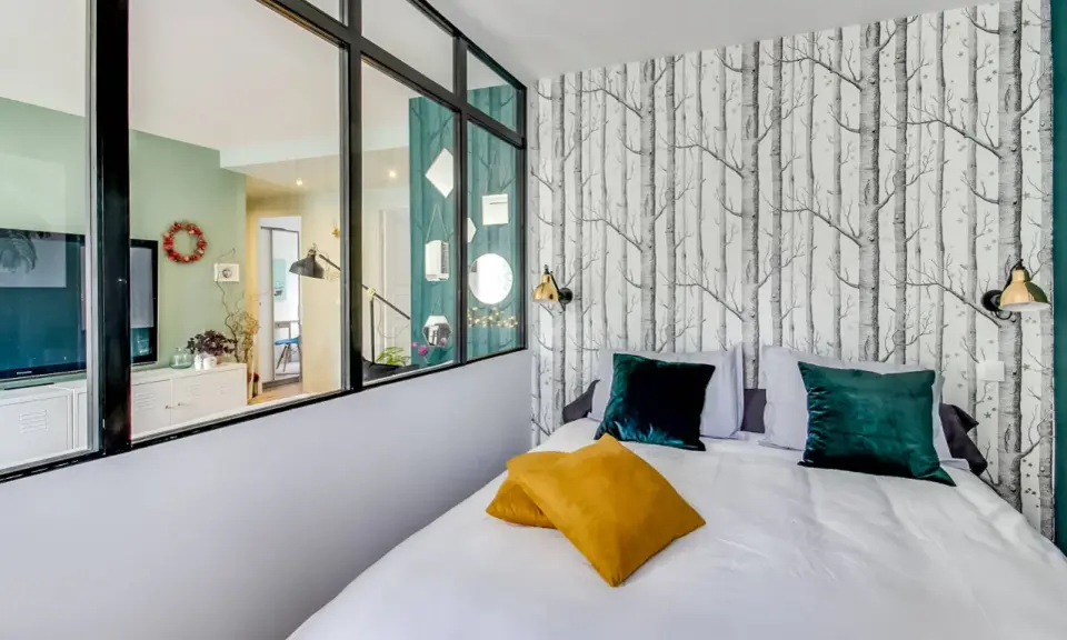 Idée pour comment amenager une chambre en longueur : un lit confortable avec des coussins colorés, un papier peint forêt et une verrière pour séparer les espaces.