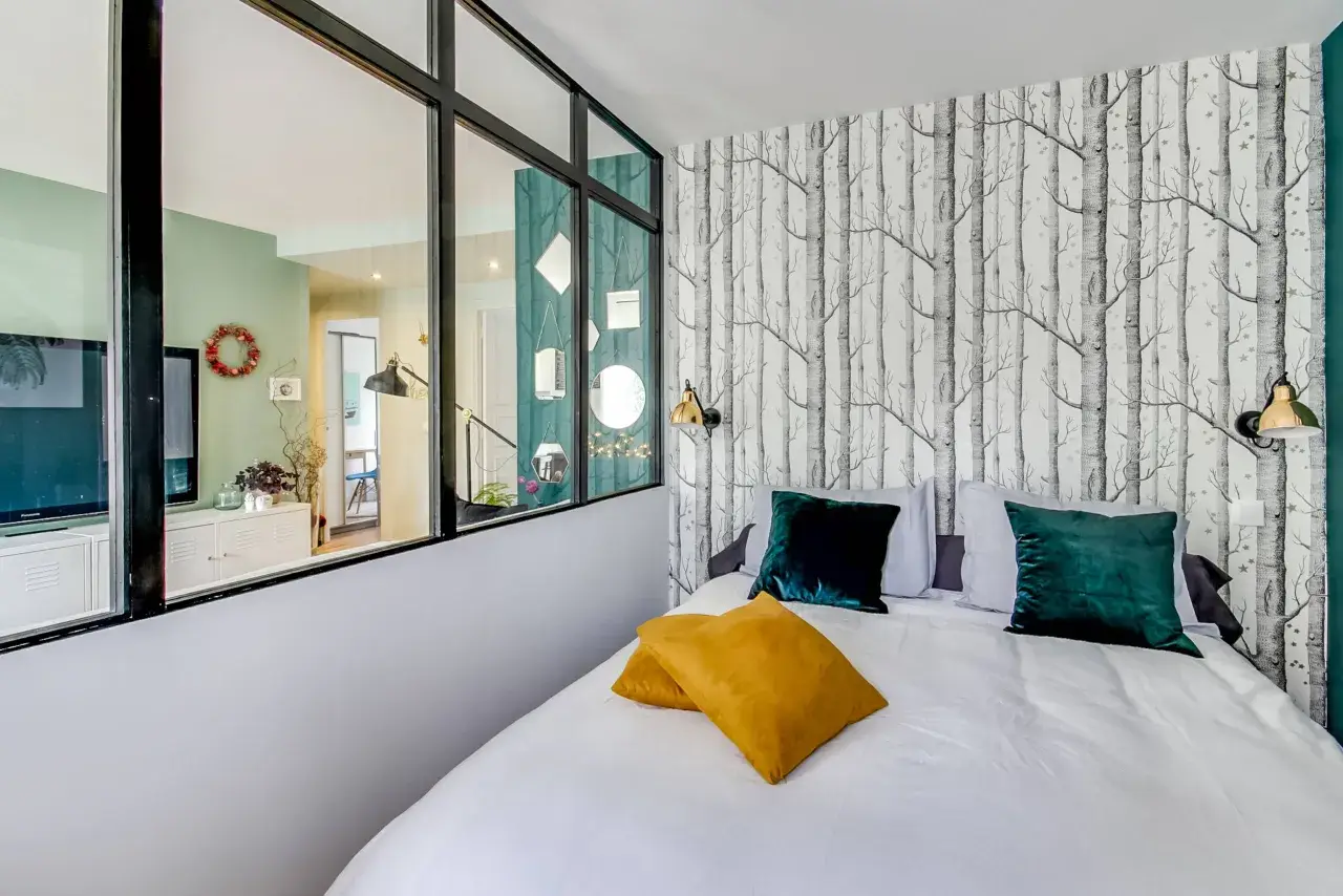 Idée pour comment amenager une chambre en longueur : un lit confortable avec des coussins colorés, un papier peint forêt et une verrière pour séparer les espaces.