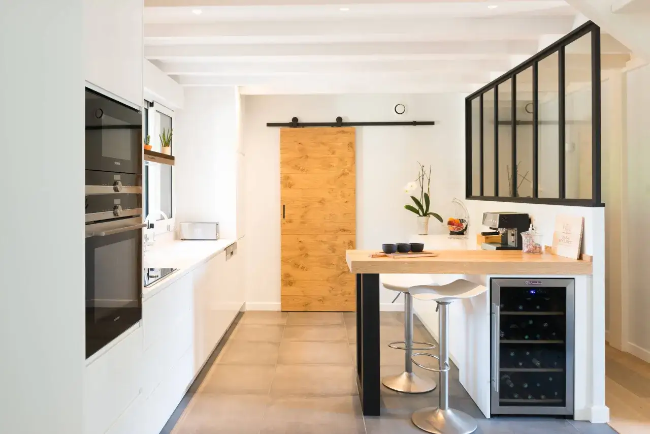 Cuisine 10m2 ferm&eacute;e, moderne et lumineuse avec porte coulissante en bois, &icirc;lot central avec cave &agrave; vin et tabourets.