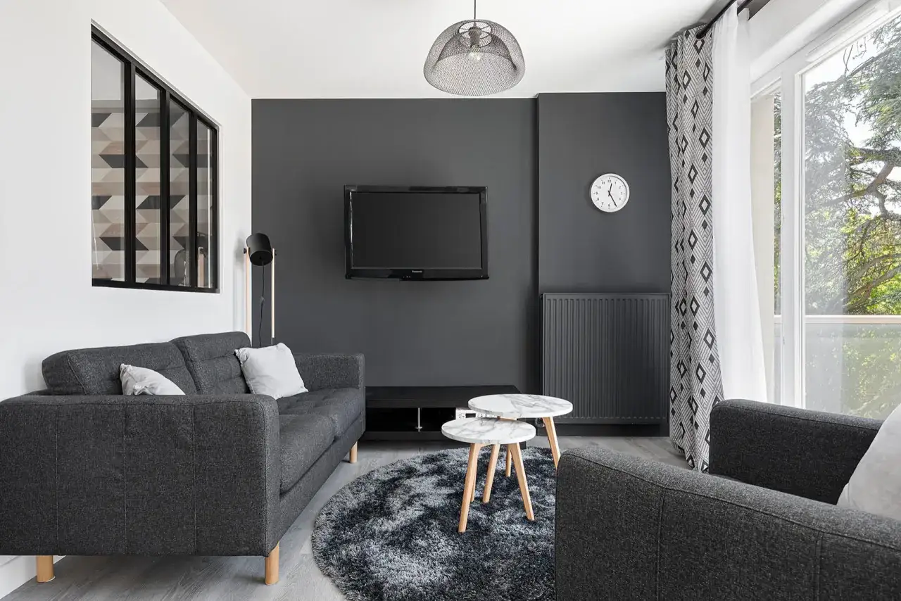 Un petit espace chambre salon moderne avec canap&eacute; gris, table basse marbre, tapis rond et TV murale.