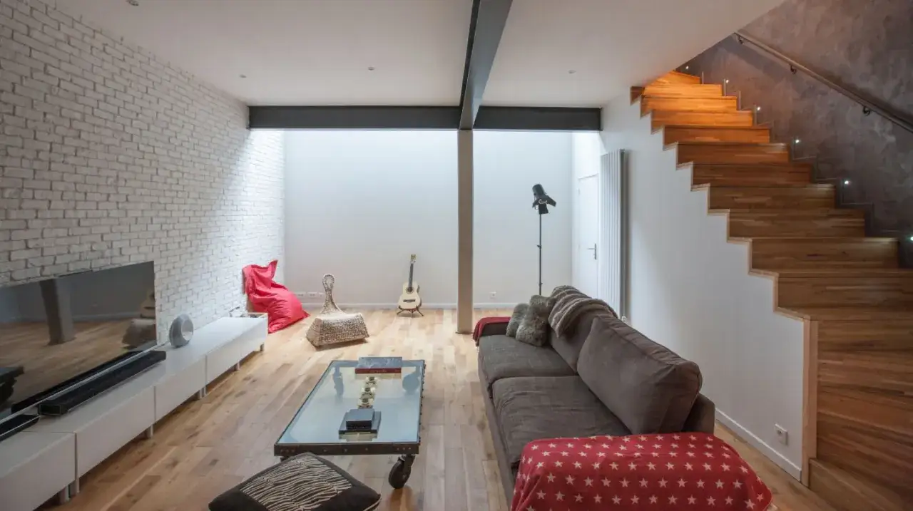 Am&eacute;nager un sous-sol en pi&egrave;ce habitable : salon moderne avec mur de briques, escalier en bois et mobilier confortable.