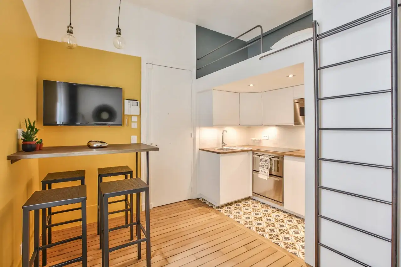 Aménagement studio 18m² : cuisine moderne, coin repas jaune vif avec TV, et lit mezzanine accessible par échelle.