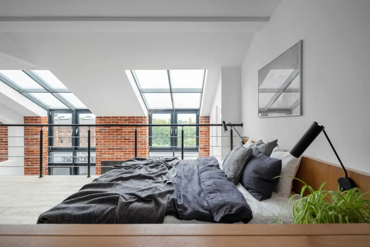 Lit douillet dans une chambre mezzanine avec vue sur des briques apparentes et un ciel lumineux par les fen&ecirc;tres de toit.