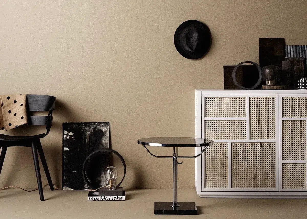 Un fauteuil noir avec un plaid &agrave; pois, une lampe sur des livres, une table ronde et une commode en rotin sur un mur couleur brun.