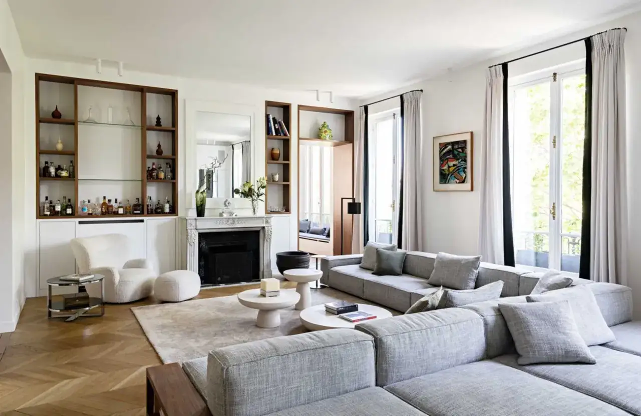 Salon lumineux avec un grand canap&eacute; gris, une chemin&eacute;e, des &eacute;tag&egrave;res remplies de bouteilles et un fauteuil blanc. La maison de r&ecirc;ve parfaite pour se d&eacute;tendre.