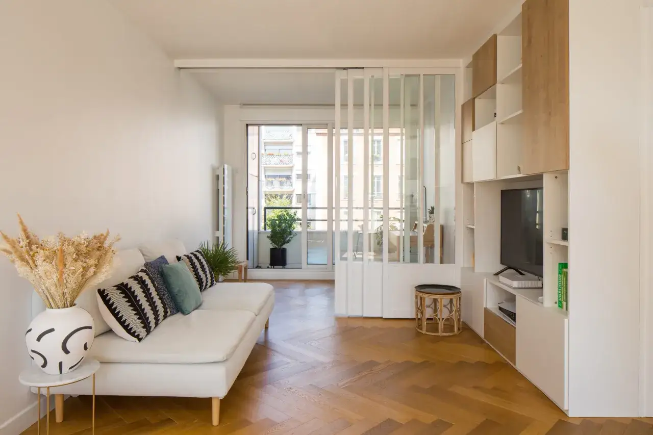 Un salon lumineux avec un canap&eacute; blanc, des coussins graphiques et un bouquet de fleurs s&eacute;ch&eacute;es. Un parquet en chevrons et une verri&egrave;re s&eacute;parent l'espace, typique d'un appartement traversant.