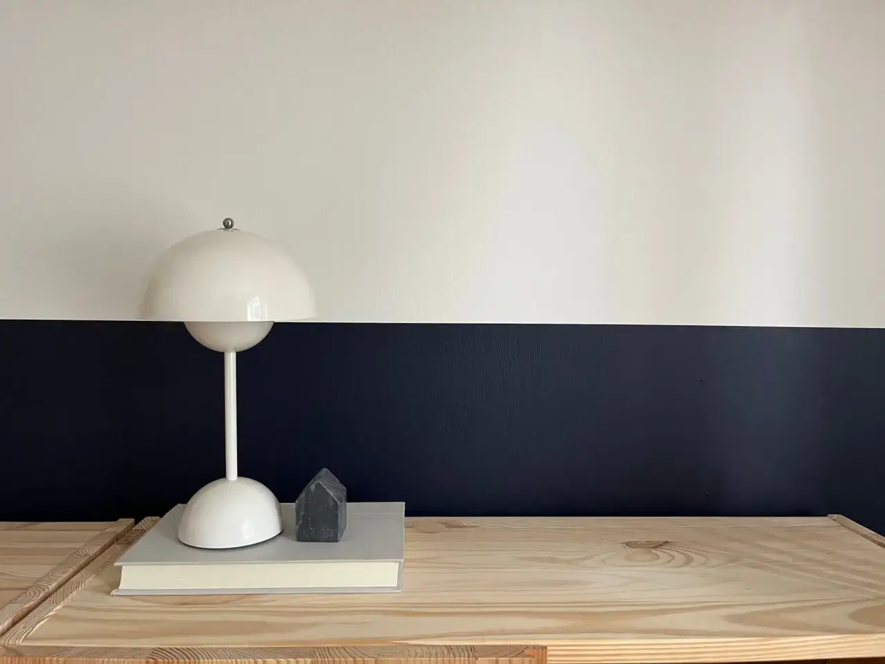Lampe blanche sur un livre, devant un mur deux couleurs diff&eacute;rentes : blanc et bleu nuit. Un petit objet gris en forme de maison est pos&eacute; sur le livre.