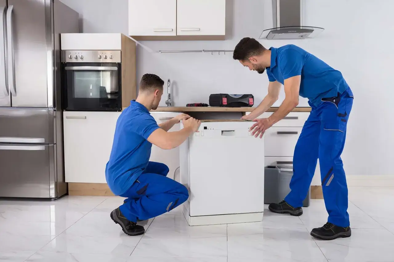 Deux techniciens en bleu installent un lave-vaisselle blanc. On peut coucher un lave-vaisselle pour le transporter, mais pas pour le faire fonctionner.