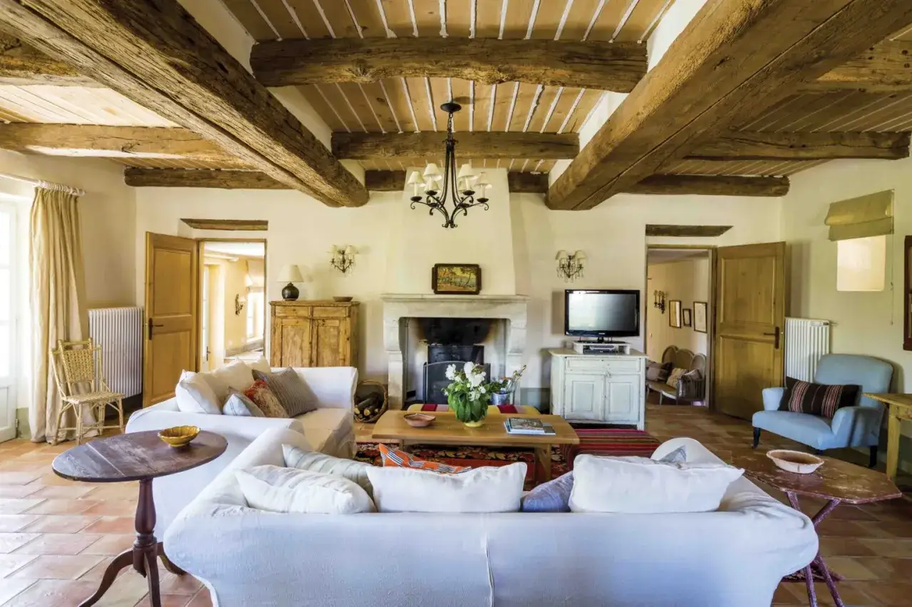 Salon d'une maison proven&ccedil;ale avec poutres apparentes, chemin&eacute;e en pierre, canap&eacute;s blancs confortables et sol en terre cuite.