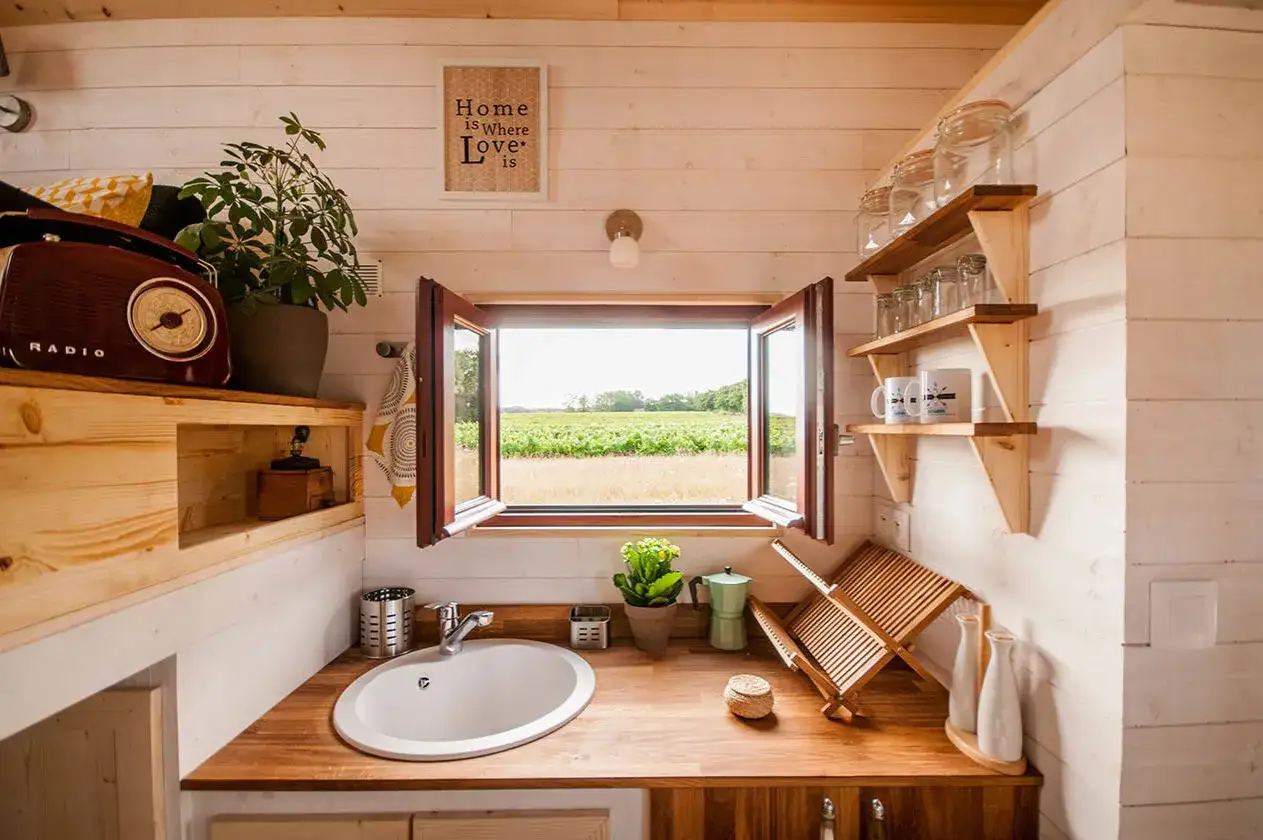 Am&eacute;nagement petite maison : cuisine fonctionnelle avec &eacute;vier, &eacute;tag&egrave;res en bois, radio vintage et vue sur la campagne.