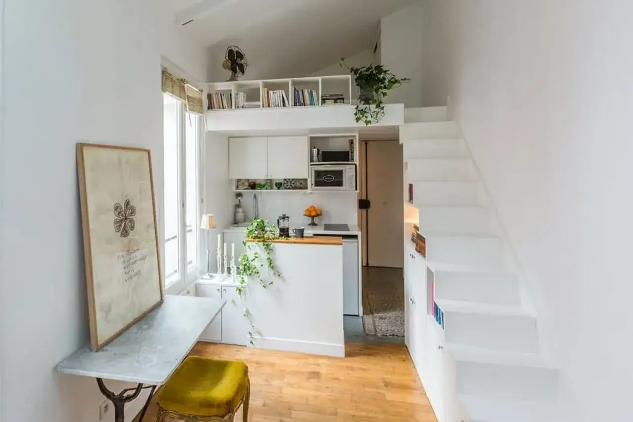 Un petit appartement optimis&eacute; avec une cuisine, un coin repas et un lit en mezzanine accessible par des escaliers blancs.