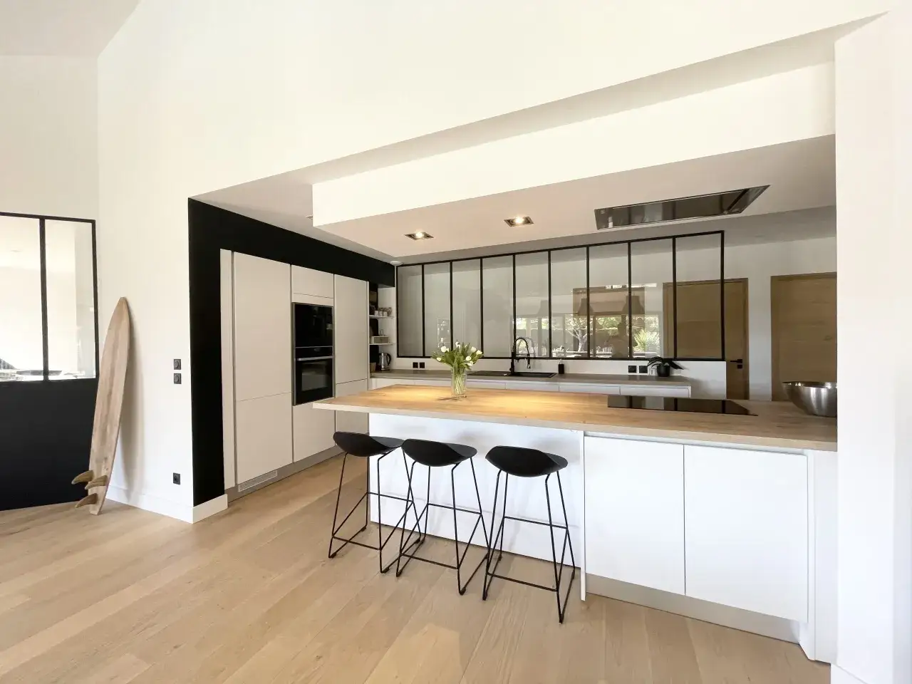 Cuisine ouverte sur salon, moderne et épurée, avec îlot central, tabourets noirs et plancher en bois clair.