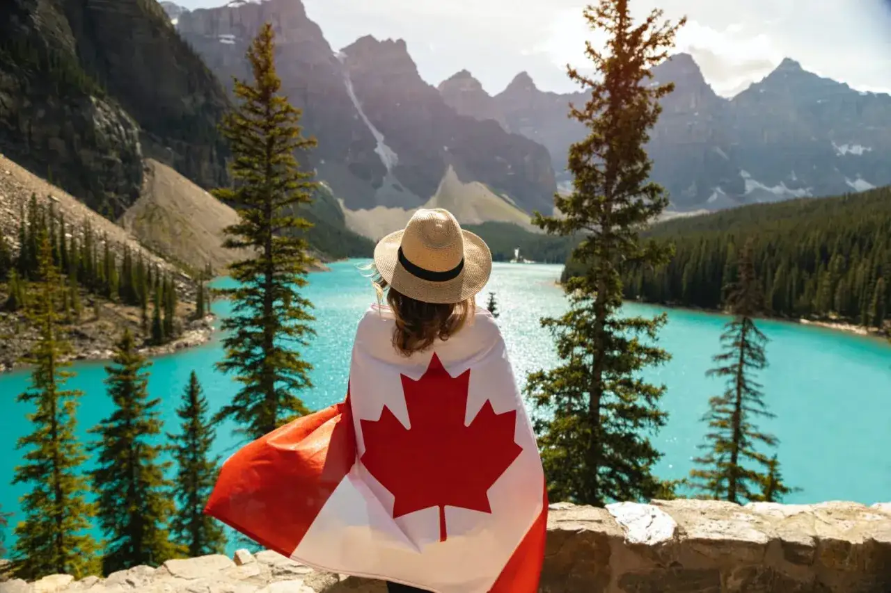 Une femme, drap&eacute;e dans le drapeau canadien, admire un lac turquoise et des montagnes majestueuses. Un r&ecirc;ve de d&eacute;m&eacute;nager au Canada.