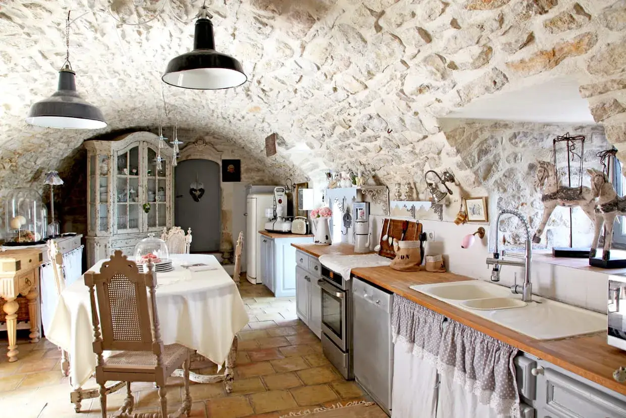 Cuisine rustique avec des vieux meubles patin&eacute;s, murs en pierre, vaisselle ancienne et une table dress&eacute;e.