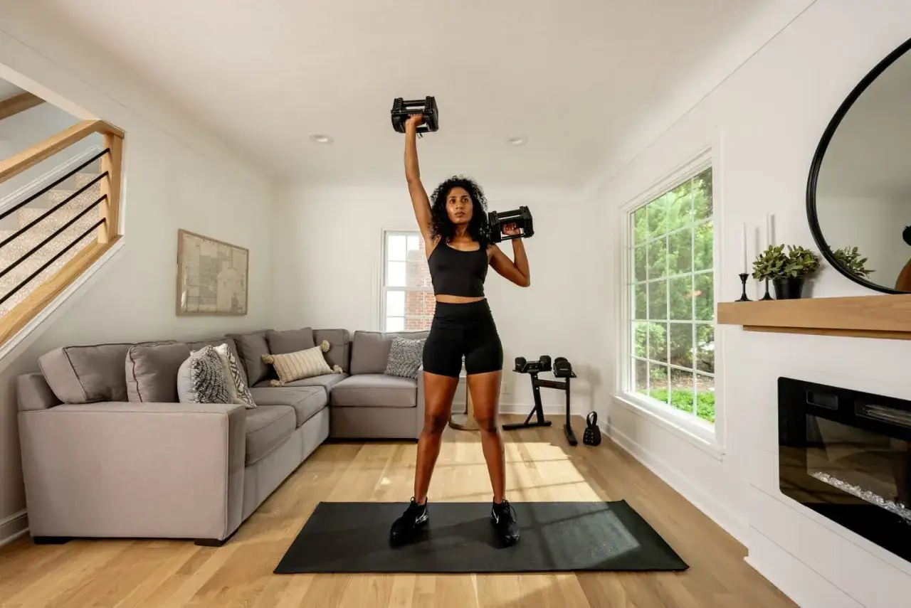 Femme faisant des halt&egrave;res pour faire une salle de sport chez soi. Un tapis de yoga est au sol.