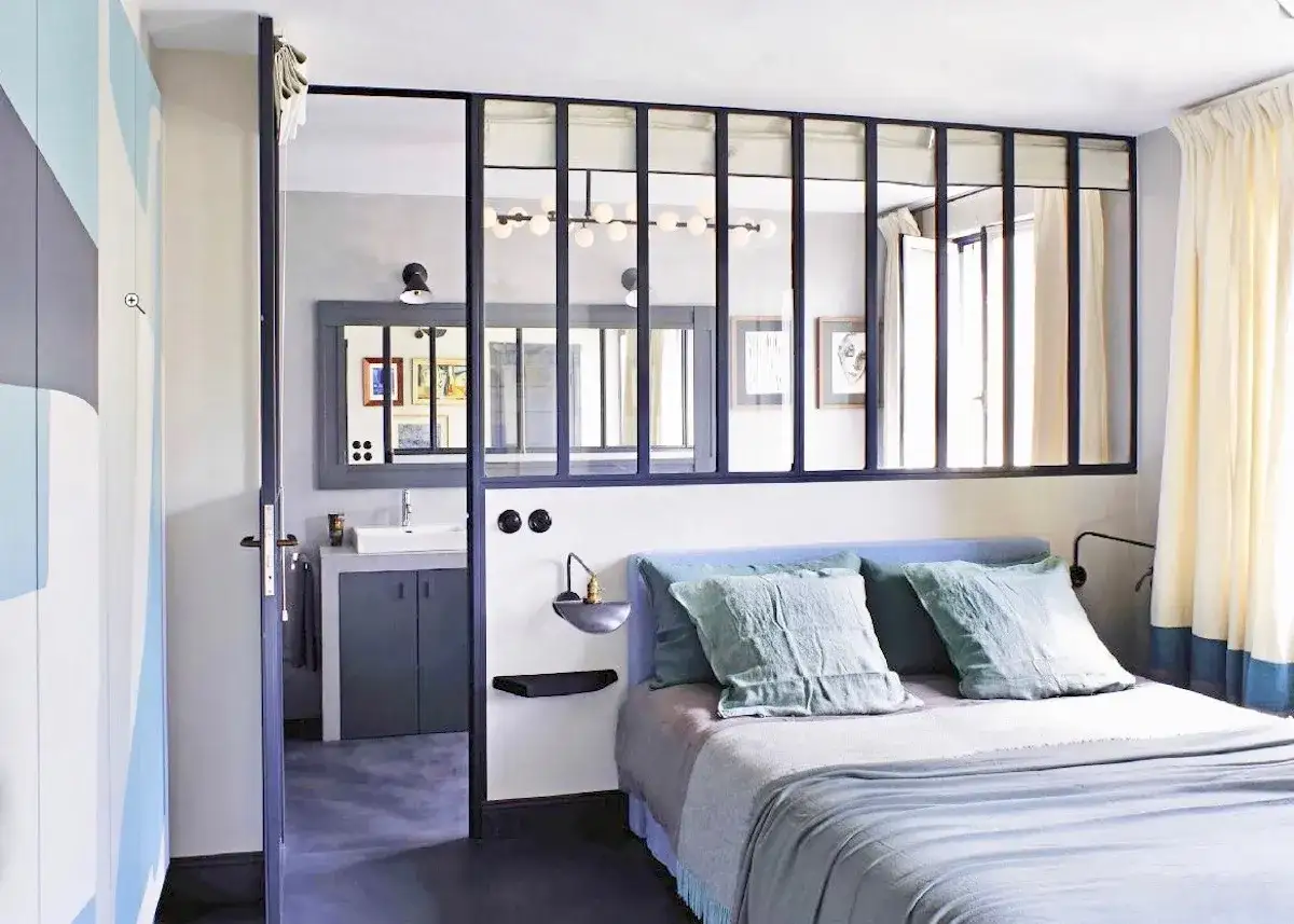 Une chambre moderne avec une verrière séparant la salle de bain. Le lit bleu est prêt pour une nuit paisible dans cette suite parentale.