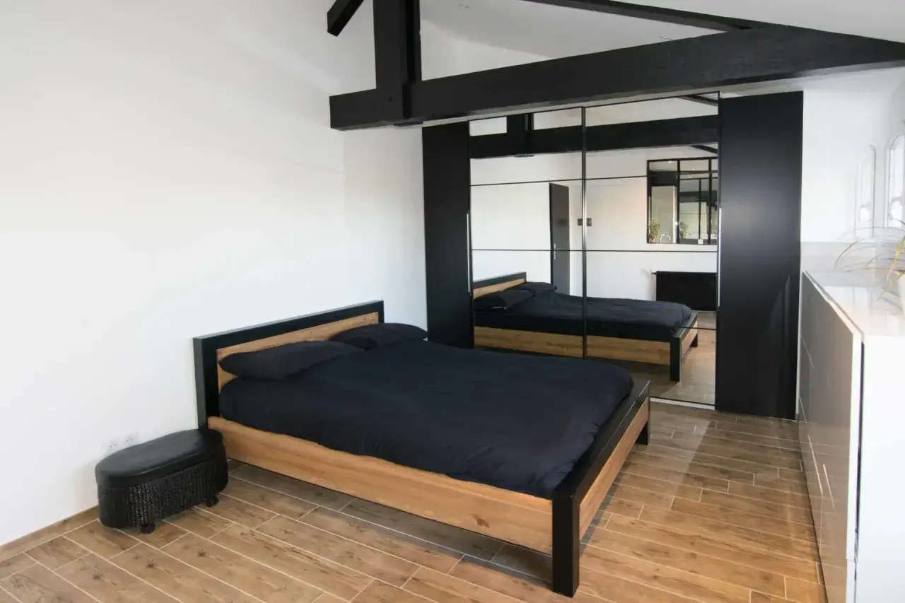 Une chambre moderne avec un lit en bois et des draps noirs, des poutres apparentes et une grande armoire miroir. Id&eacute;al pour refaire sa chambre avec style.