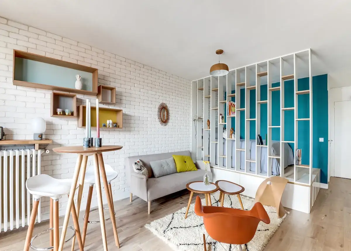 Am&eacute;nagement studio 25 m2 : salon cosy avec canap&eacute; gris, table haute, &eacute;tag&egrave;res murales et un lit s&eacute;par&eacute; par une biblioth&egrave;que ajour&eacute;e.