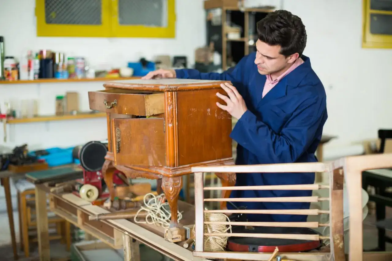 Un artisan examine un meuble en bois, pr&ecirc;t &agrave; le repeindre. Des outils et des mat&eacute;riaux sont visibles dans l'atelier.